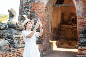 glad turistkvinna i vit klänning tar foto med mobil smartphone, under besök i wat chaiwatthanaram tempel i ayutthaya historiska park, sommar, solo, asien och thailand resekoncept