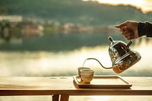 hand som håller vintage tekanna och häller varmt te till koppen på träbord mot sjöutsiktsbakgrund på kaféet i morgonsoluppgången, ban rak thai village, mae hong son provinsen, thailand foto