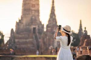 glad turistkvinna i vit klänning tar foto med mobil smartphone, under besök i wat chaiwatthanaram tempel i ayutthaya historiska park, sommar, solo, asien och thailand resekoncept