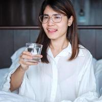 glad kvinna håller vattenglas, kvinna dricker rent vatten på sängen hemma. hälsosam, förfriskning, livsstilskoncept foto