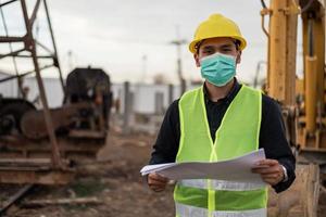 ingenjör man bär ansiktsmask och arbetar på byggarbetsplatsen. asiatisk arbetare man står och korsade armar på byggarbetsplatsen. foto