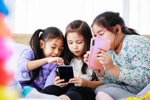 tre asiatiska söta små flickor som spelar smartphone för att spela eller studera tillsammans hemma foto