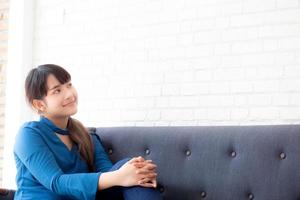 vacker ung asiatisk kvinna som sitter och ler glad och ser något på soffan som visar framtid eller planering med budskap om dig, flicka glad och koppla av på soffan hemma, livsstilskoncept. foto