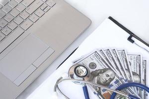 högvinkelvy av stetoskop och dollarsedlar på urklipp med bärbar datortangentbord på vit marmorskiva, medicinsk kostnad och försäkringskoncept foto