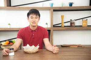 asiatisk syoung man med casual röd t-shirt njuta av att äta frukost, äta pommes frites. ung man lagar mat i köksrummet i loftstil foto