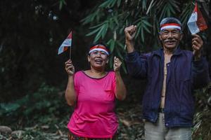 indonesiska äldre par som firar Indonesiens självständighetsdag tillsammans i byn. gammal man och kvinna med rött och vitt pannband och håller den lilla indonesiska flaggan. foto