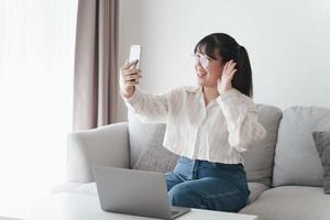 ung asiatisk kvinna som använder smartphone för online videokonferenssamtal viftande hand gör hej gest på soffan i vardagsrummet. foto