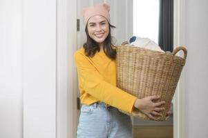ung glad kvinna som bär gul skjorta som håller en korg full av kläder hemma, tvättkoncept foto