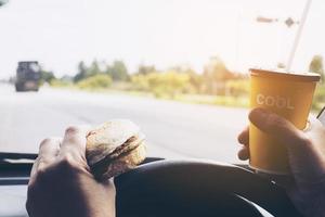 man kör bil medan han håller en kopp kallt kaffe och äter hamburgare foto