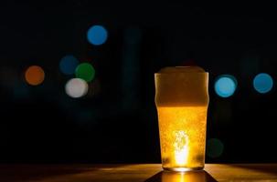 ett glas öl med dess skum sätter på träbord av baren isolerad på mörk natt bakgrund med färgglada bokeh lampor på takbaren. foto