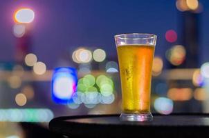 ett glas öl sätter på bordet på takbaren med färgglad stadsbokeh ljus bakgrund. foto