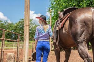 vacker blond cowgirl med hatt som står baklänges nära hästranchbakgrunden foto