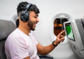 passagerare i flygplan som rör vid lcd-underhållningsskärm. latinamerikansk man i flygkabin med smart enhet som lyssnar på musik i hörlurar. foto