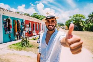 turist gör selfie i porto seguros historiska centrum. latinamerikansk man i hatt och tummen upp ler mot kameran foto