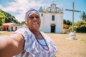 glad brasiliansk kvinna av afrikansk härkomst klädd i den traditionella bahianska klänningen och gör en selfie framför kyrkan. fokusera på ansiktet foto