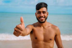 atletisk ung latinamerikansk man på stranden med tummen upp. leende man tittar på kameran foto