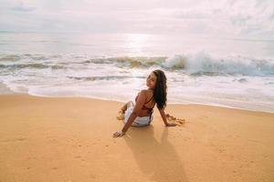 siluett av ung kvinna på stranden. latinamerikansk kvinna sitter på stranden sanden tittar på himlen en vacker sommardag foto