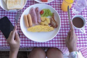 färskt frukostbord med omelettrulle, korv, bacon, skinka, tomat och potatis på ett fat. kvinnans händer som håller gaffel och kniv ingår kaffe apelsinjuice, bröd och mobiltelefon på ett bord. foto