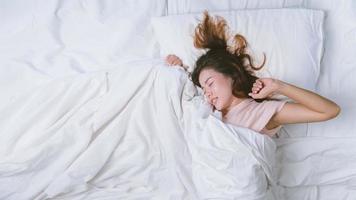 ung kvinna sover gott i sängen kramar mjuk vit kudde. tonårsflicka vilar. god natt sömn koncept. flicka som bär en pyjamas sover på en säng i ett vitt rum på morgonen. varm ton. foto