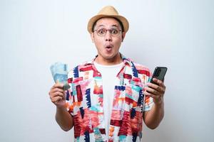 Asiatisk ung man som visar wow ansiktsuttryck medan han håller mobiltelefon och papperspengar foto