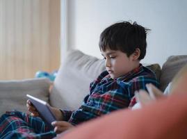 barn som sitter i soffan och tittar på tecknad film, ung pojke spelar spel online på surfplatta med vänner på semester, barnpojke bär pyjamas och kopplar av hemma på morgonen foto