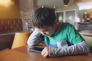 filmiskt porträtt ung pojke spelar spel på mobilen medan han väntar på mat, barn som sitter på kaféet och skickar text till vänner, barn spelar spel online på telefonen. barn med teknikkoncept foto