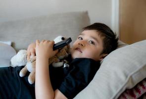 porträtt ung pojke som håller fjärrkontrollen och tittar upp med ett nyfiket ansikte, barn som sitter i soffan och tittar på tecknad film på tv, barn ligger ner på soffan och kopplar av i vardagsrummet efter hemkomsten från skolan. foto