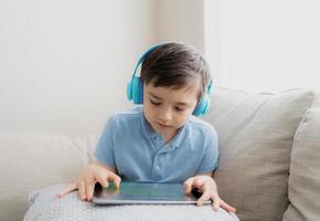 glad ung pojke som bär hörlurar för att spela spel på internet med vänner, barn som sitter bredvid fönstret och läser eller tittar på tecknad film på surfplattan, skolbarn gör läxor online hemma. foto