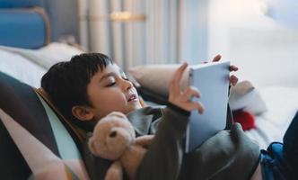glad unge som ligger i sängen och håller surfplattan tittar på tecknad film och chattar med vänner på digital block, söt ung pojke spelar spel online på internet, barn kopplar av hemma med familjen foto