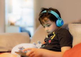 glad ung pojke som bär hörlurar för att spela spel online på internet med vänner, barn som sitter i soffan och läser eller tittar på tecknad film på surfplattan barn kopplar av hemma på morgonen på helgen foto