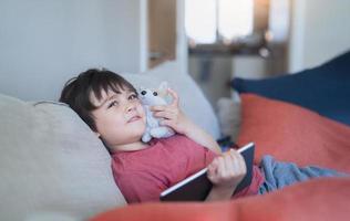 glad unge som sitter på soffan och leker med hundleksak och tittar på tecknade filmer eller spelar spel på surfplatta, barnpojke kopplar av efter att ha lärt sig online på internet, hemundervisning eller onlineutbildningskoncept foto