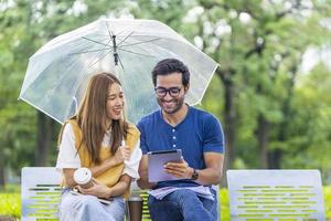 unga asiatiska och indiska par njuter av att koppla av i regnet tillsammans i den offentliga parken medan de sitter tillsammans på bänken under helgen foto