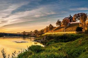 antik ortodox kyrka på den höga stranden framför floden vid vacker solnedgång foto