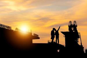 siluett av ingenjör och arbetare med urklippsbana som kontrollerar projekt på byggarbetsplatsbakgrund, infrastrukturbyggarbetsplats vid solnedgången på kvällstid foto