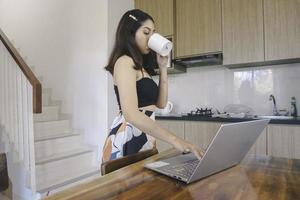 ung asiatisk kvinna tillbringar sin tid hemma och arbetar på distans i matsalen med en onlinekonferens på sin bärbara dator medan hon bär en mugg. foto