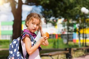 flicka med en ryggsäck ta av sig den medicinska masken och äta paj nära skolan. ett snabbt mellanmål med en bulle, onyttig mat, lunch hemifrån. tillbaka till skolan. utbildning, grundskoleklasser, 1 september foto