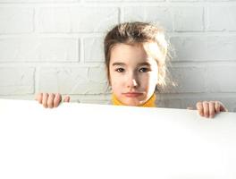 en ledsen tjej håller ett vitt pappersark - mock-up för reklam, slogan, inskription. kopieringsutrymme är i barns händer, barnet är upprört och gömt. foto
