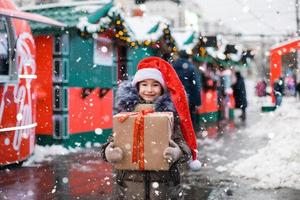 porträtt av glad flicka i tomtehatt med presentförpackning till jul på stadsgatan på vintern med snö på festlig marknad med dekorationer och fairy lights. varma kläder, stickad halsduk och päls. nyår foto