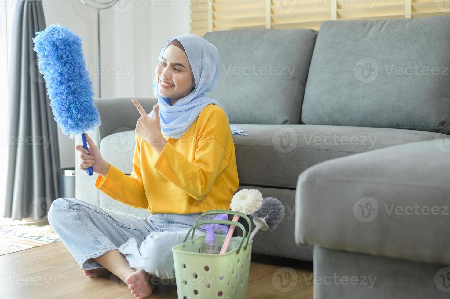 jonge gelukkige moslimvrouw die gele handschoenen draagt en een mand met schoonmaakmiddelen in de woonkamer vasthoudt. foto