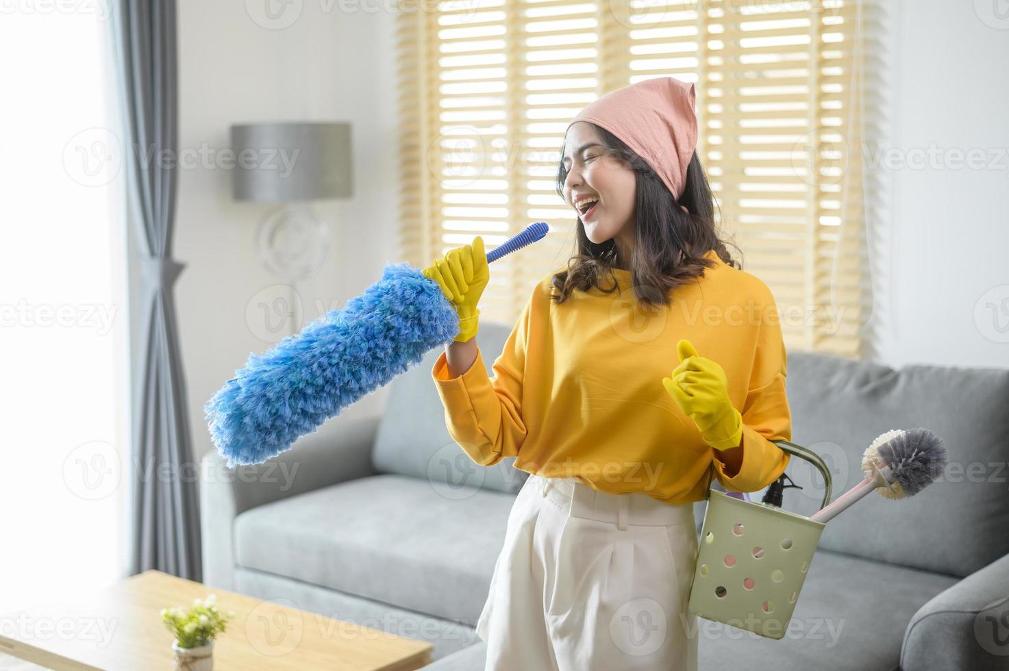 jonge gelukkige vrouw die gele handschoenen draagt en een mand met schoonmaakmiddelen in de woonkamer vasthoudt. foto