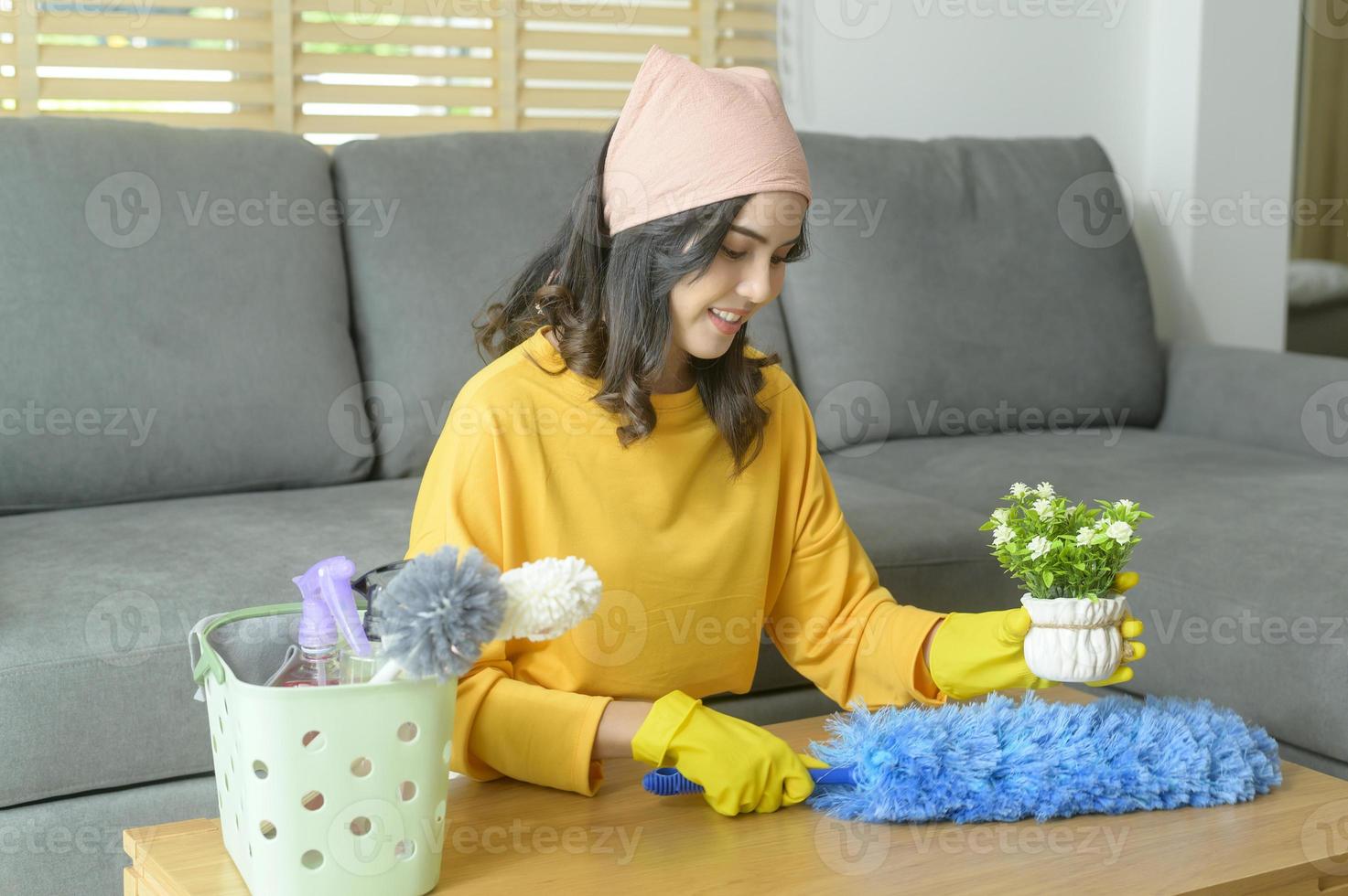 jonge gelukkige vrouw die gele handschoenen draagt en de tafel in de woonkamer afstoffen. foto