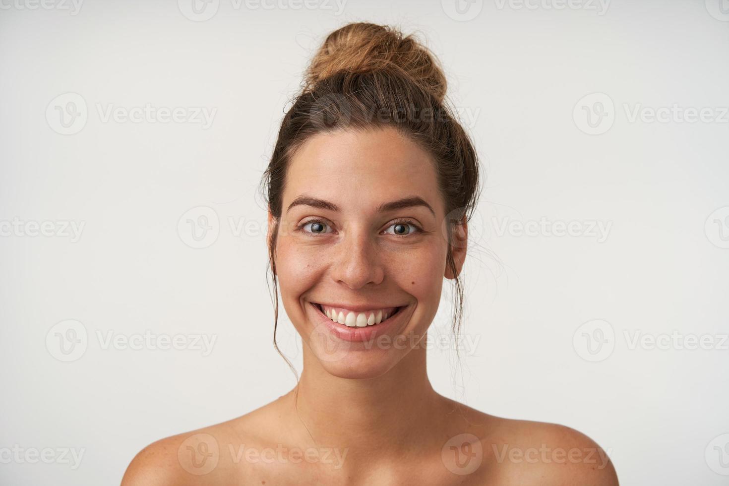 Gelukkige mooie jonge vrouw zonder make-up die op een witte achtergrond staat, vrolijk lacht naar de camera met een hoog broodje op het hoofd, concept van positieve emoties foto