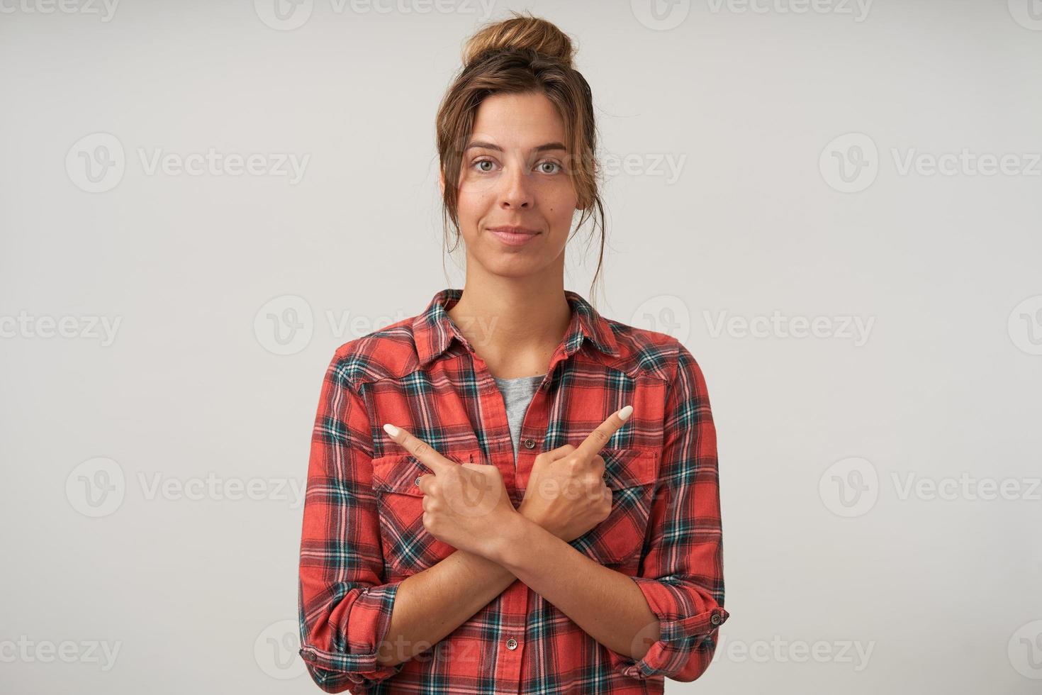 Jonge aantrekkelijke vrouw die zich voordeed op een witte achtergrond met gekruiste handen op haar borst en met wijsvingers naar verschillende kanten wijst, kijkend naar de camera met kalm gezicht foto