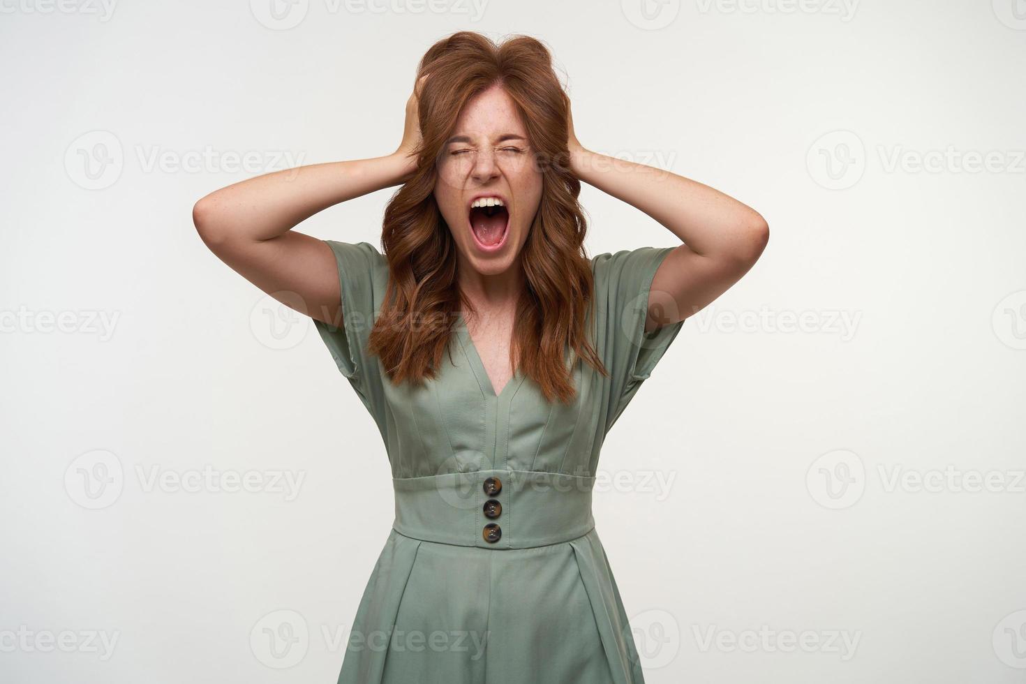 gestresste aantrekkelijke jonge vrouw met rood haar die zich voordeed op een witte achtergrond, hoofd met handen vasthoudt en met gesloten ogen schreeuwt, slechte dag foto