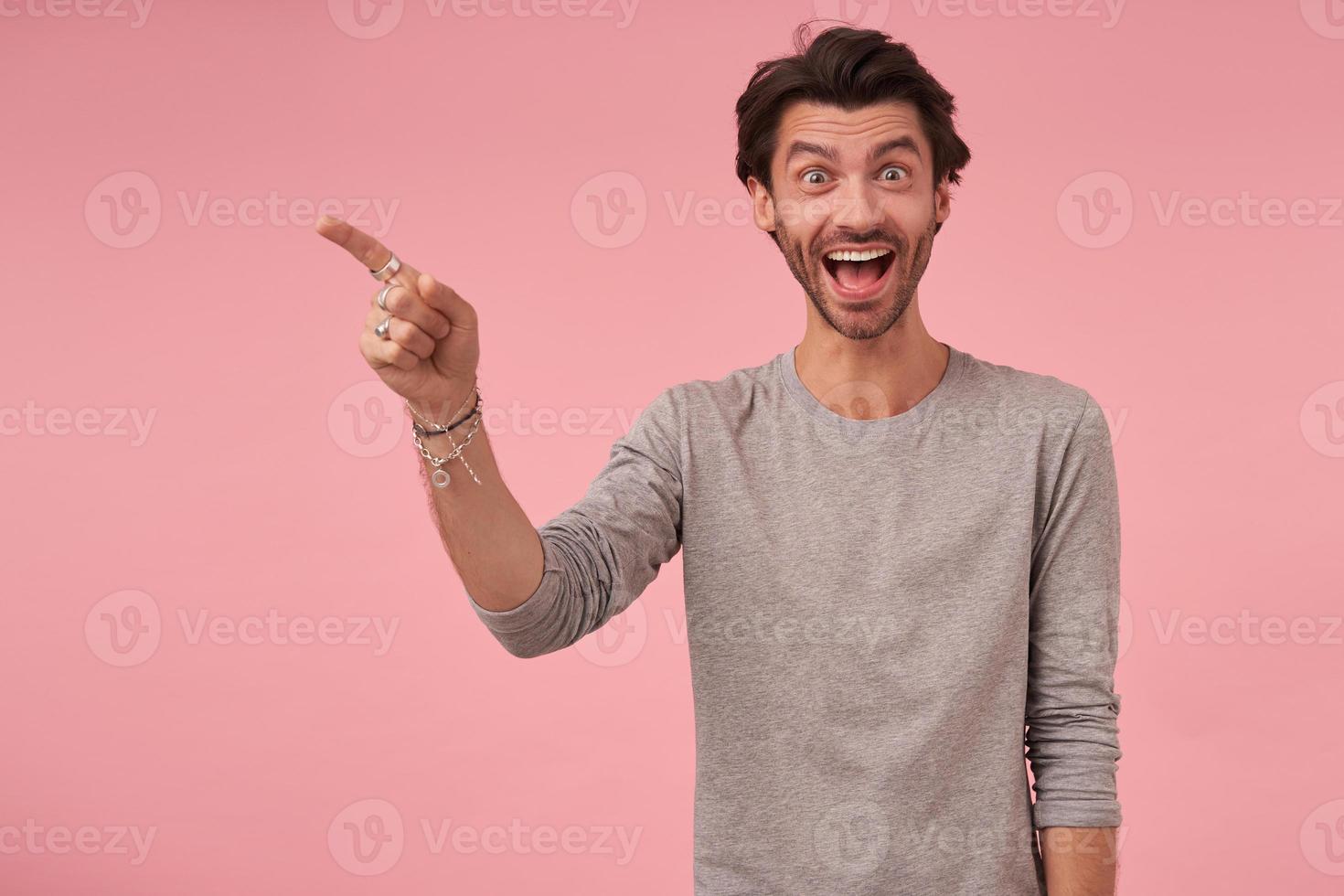 studio-opname van een vrolijke bebaarde man met trendy kapsel die over een roze achtergrond staat, casual kleding draagt, verbaasd naar de camera kijkt en wenkbrauwen optrekt, opzij wijzend met wijsvinger foto