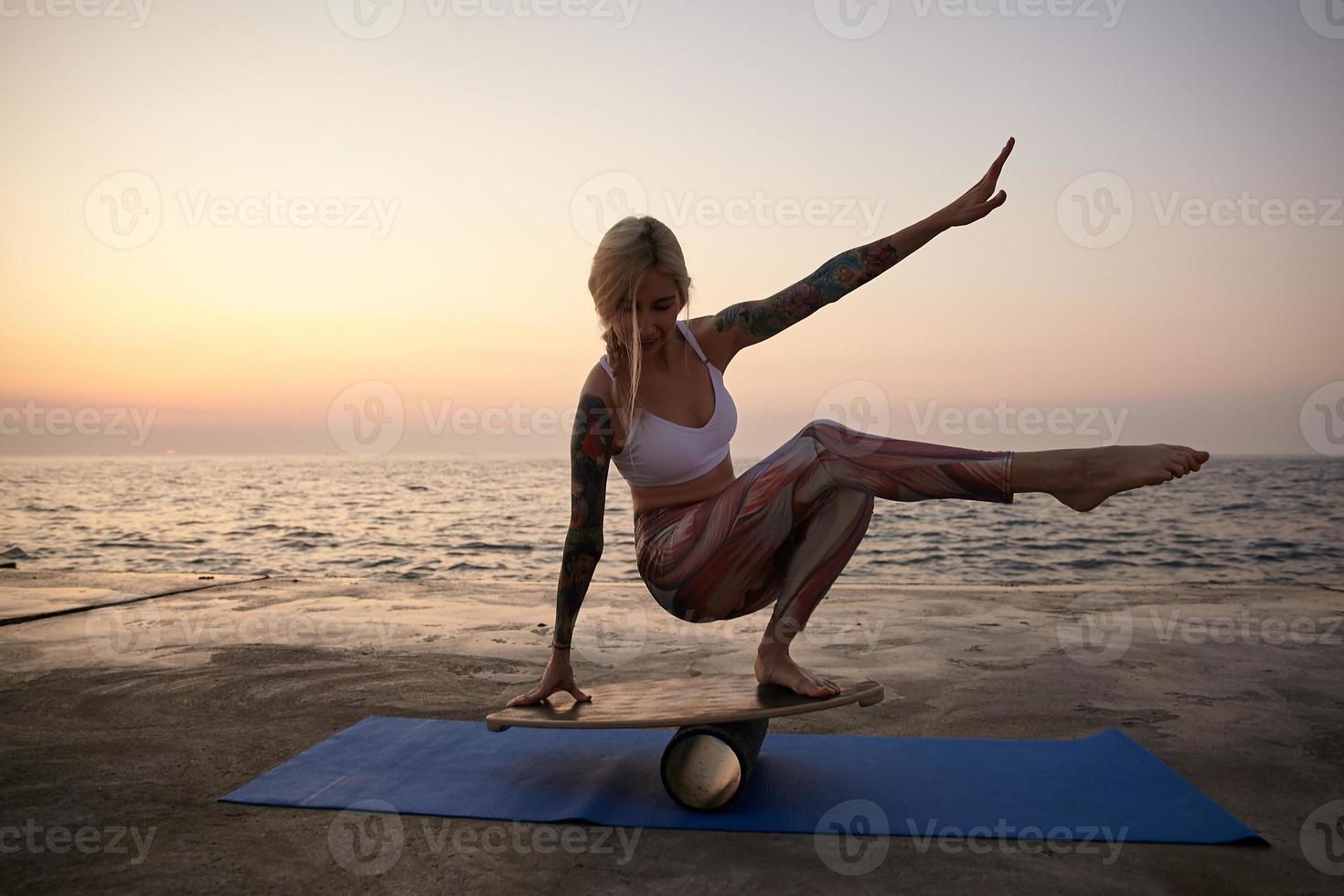 buitenopname van jonge sportieve getatoeëerde vrouw met casual kapsel die haar evenwicht houdt op de sportuitrusting, poseren aan de kust tijdens zonsopgang, sportieve kleding dragen foto