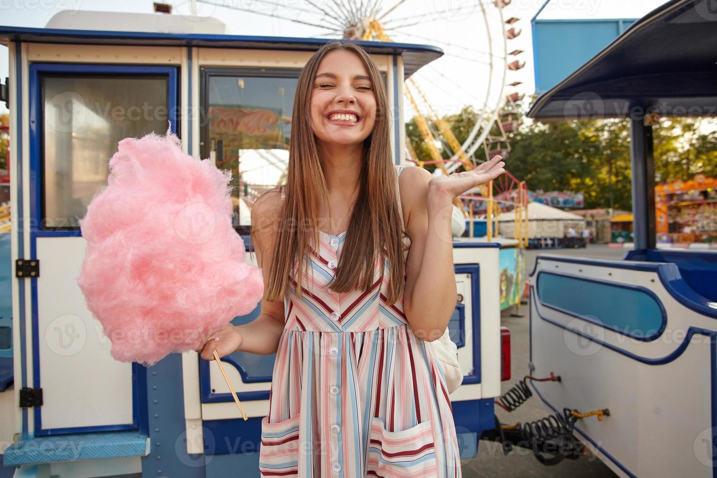 Positieve jonge mooie brunette vrouw met lang haar poseren over pretpark, staande met roze suikerspin in de hand en gesloten ogen, palm opsteken en vrolijk glimlachen foto