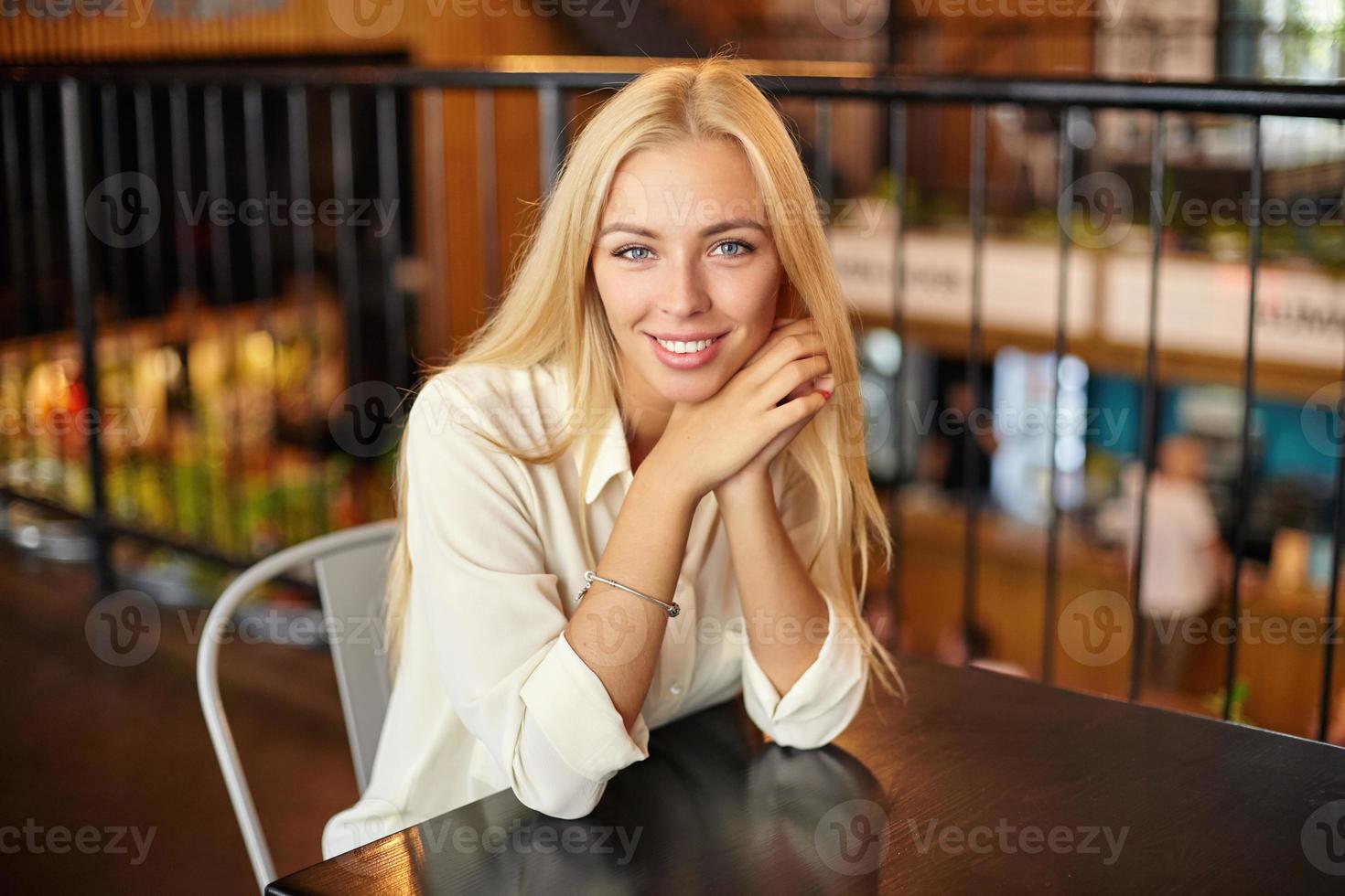 indoor portret van mooie jonge langharige blonde vrouw poseren over café interieur, camera kijken met charmante glimlach en leunend hoofd op gekruiste armen foto