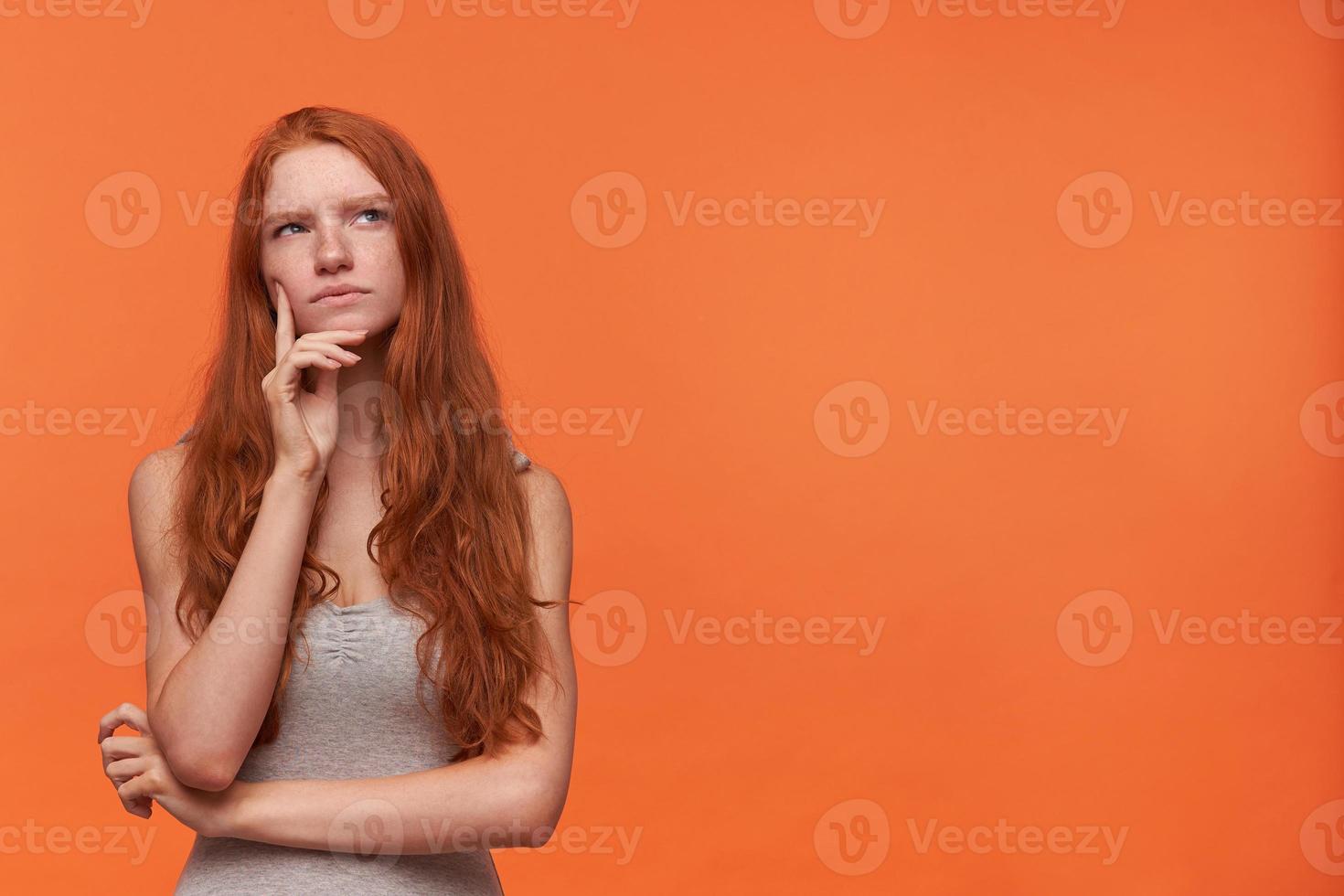 schattige jonge peinzende vrouw met golvend foxy haar lond haar naar boven kijkend terwijl ze denken, geïsoleerd over oranje achtergrond, casual grijs shirt dragend, hand op haar gezicht houdend foto