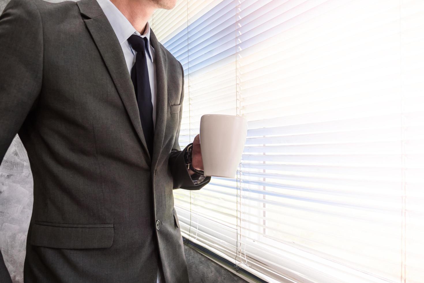 close-up van zakenman die een koffiekopje vasthoudt terwijl hij voor een raam in een kantoor staat foto